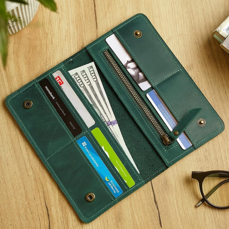 May include: Open, emerald green leather wallet with multiple card slots, a zippered compartment, and snap closures. The wallet contains several credit cards and US $100 bills. The wallet is open on a wooden surface.