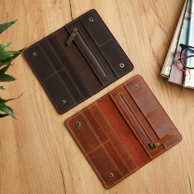 May include: Two open leather wallets in shades of brown and tan are displayed on a wooden surface. Each wallet features a zippered compartment and multiple card slots. The wallets are rectangular and have snap closures.