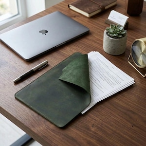 May include: A dark green leather document folder with a folded corner, containing a stack of white papers. The folder is placed on a wooden surface alongside a silver laptop, a metal pen, a small potted succulent, and a gold desk clock.
