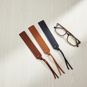 May include: Three leather bookmarks in brown, tan, and navy blue, each with a matching tassel. A pair of eyeglasses with a brown frame rests nearby on a light wood surface. The bookmarks are rectangular with rounded ends.