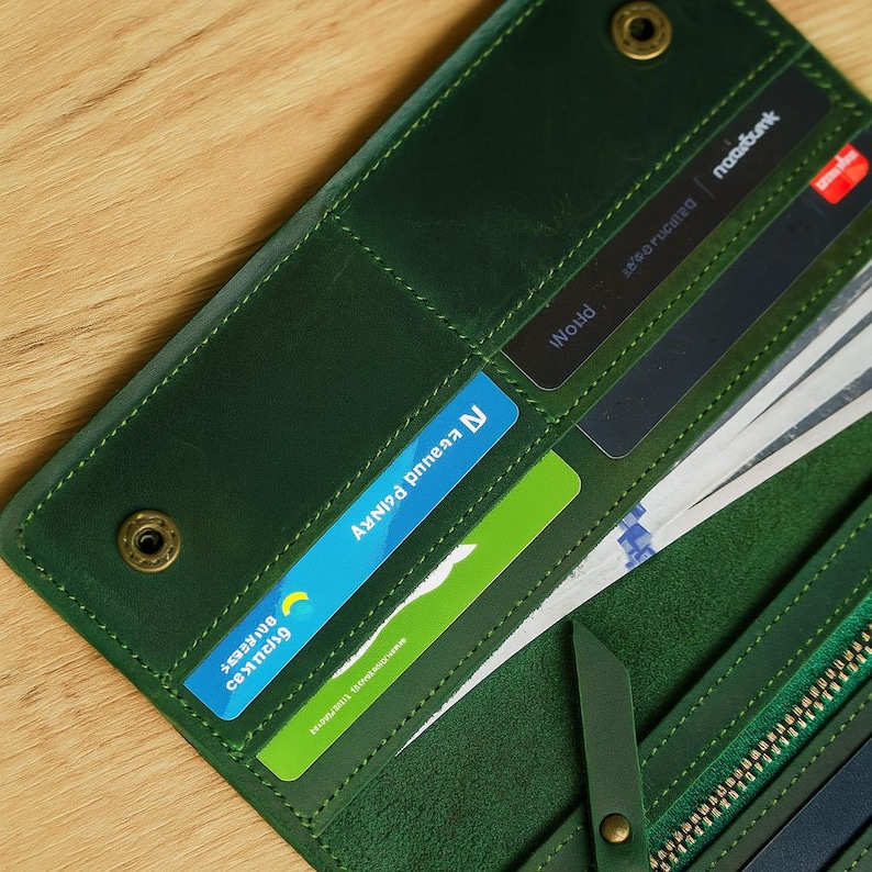 May include: A green leather wallet, open to show credit cards and cash. The wallet has a zip closure and a snap closure. The stitching is a lighter green. The wallet is on a wooden surface.