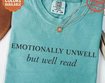 May include: Seafoam green t-shirt with the text "Emotionally Unwell but well read" in black. The shirt has a crew neck and short sleeves. A gold necklace with a pendant is draped on the shirt. The shirt is labeled "Comfort Colors".