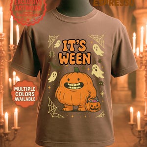Puede incluir: Camiseta marrón con un diseño de Halloween. El diseño presenta una calabaza naranja sonriente sosteniendo un cubo de caramelos, rodeada de fantasmas, arañas y el texto "IT'S WEEN". La camiseta tiene cuello redondo y mangas cortas.