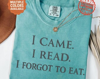 May include: Seafoam green t-shirt with the text "I CAME. I READ. I FORGOT TO EAT." A gold necklace with a pendant is on the shirt. The shirt has a Comfort Colors tag and a "Multiple Colors Available" sticker.