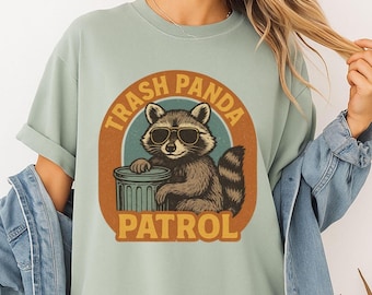 May include: A light green t-shirt featuring a graphic of a raccoon wearing sunglasses and leaning on a trash can. The text "Trash Panda Patrol" is printed in a circular design around the raccoon.