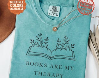 Może przedstawiać: Turkusowa koszulka z napisem "BOOKS ARE MY THERAPY" i grafiką otwartej książki. Koszulka ma okrągły dekolt i krótkie rękawy. Złoty naszyjnik z wisiorkiem jest zarzucony na koszulkę. Koszulka jest oznaczona jako "Comfort Colors".