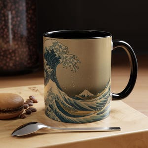 Puede incluir: Una taza de cerámica con asa e interior negros, que muestra la obra de arte japonesa "La Gran Ola de Kanagawa". El diseño muestra una gran ola, barcos y el monte Fuji. Adecuada para bebidas calientes.