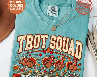 May include: A seafoam green short-sleeve t-shirt featuring a graphic of four turkeys running. The text reads TROT SQUAD and THANKSGIVING 5K. The shirt has a crew neckline and a Comfort Colors brand label on the inside collar. A thin gold chain necklace rests on the shirt.