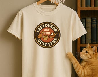 May include: Ivory-colored t-shirt with a circular graphic that reads "Leftovers are for Quitters." The graphic features a drumstick, a box, and a slice of pie. The shirt is hanging on a wooden hanger.