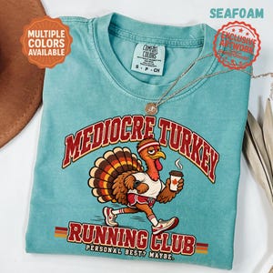 May include: A seafoam green t-shirt featuring a graphic of a turkey wearing a headband, white t-shirt, and shorts while running and holding a coffee cup. The text reads MEDIOCRE TURKEY RUNNING CLUB PERSONAL BEST? MAYBE. The shirt has a Comfort Colors label.
