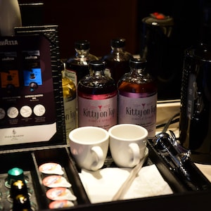 May include: A black tray with coffee supplies, including coffee pods, white mugs, and bottles of syrup labelled "Kitty on Top." A coffee maker and Lavazza coffee selection are also visible.