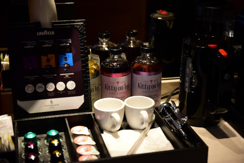 May include: A black tray with coffee supplies, including coffee pods, white mugs, and bottles of syrup labelled "Kitty on Top." A coffee maker and Lavazza coffee selection are also visible.