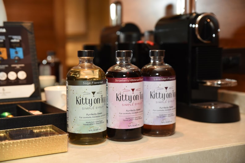 May include: Three bottles of "Kitty on Top" simple syrup in sage, strawberry hibiscus, and lavender flavours. The bottles have black caps and white labels. The bottles are in front of a coffee machine.