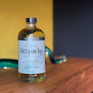 May include: A clear glass bottle of "Kitty on Top" sage-flavored simple syrup. The label reads "Purrfectly Sweet" and "Made from Cane Sugar." The bottle contains 473 ml of syrup and has a silver cap.
