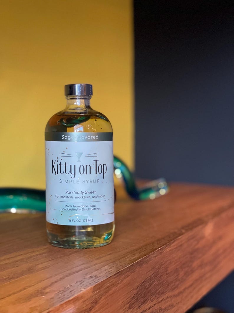 May include: A clear glass bottle of "Kitty on Top" sage-flavored simple syrup. The label reads "Purrfectly Sweet" and "Made from Cane Sugar." The bottle contains 473 ml of syrup and has a silver cap.