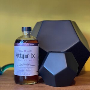 May include: A bottle of "Kitty on Top" lavender-flavored simple syrup, alongside two black geometric decorative objects. The syrup bottle is labelled "Purrfectly Sweet" and contains 473 ml.