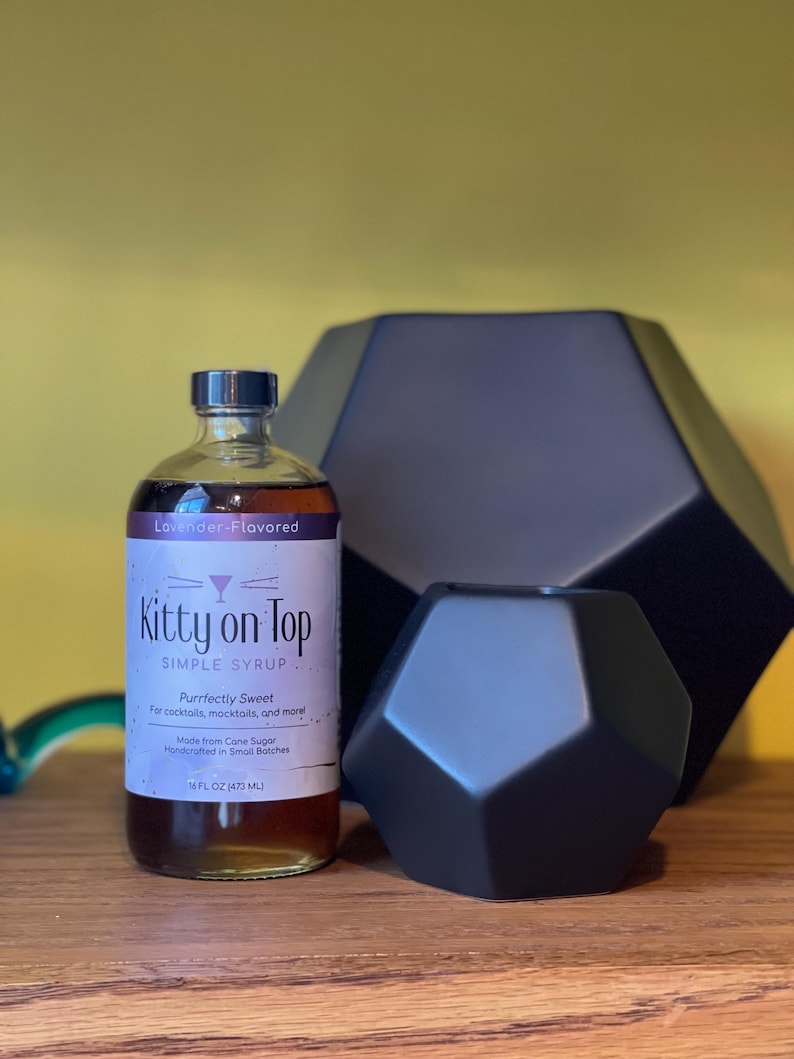May include: A bottle of "Kitty on Top" lavender-flavored simple syrup, alongside two black geometric decorative objects. The syrup bottle is labelled "Purrfectly Sweet" and contains 473 ml.
