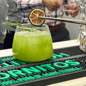 May include: A vibrant green cocktail in a clear glass, garnished with a dried citrus slice and rosemary. The glass has the text "Cullen Construction". The drink sits on a black bar mat with the word "TORNITOS" in green.
