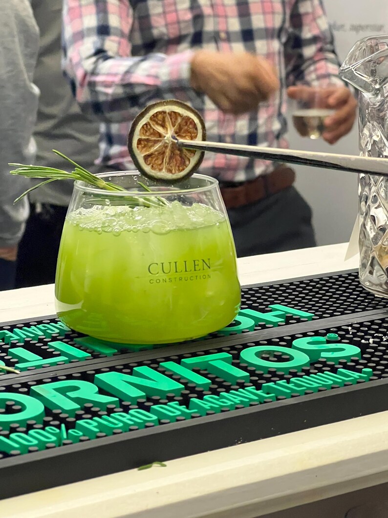 May include: A vibrant green cocktail in a clear glass, garnished with a dried citrus slice and rosemary. The glass has the text "Cullen Construction". The drink sits on a black bar mat with the word "TORNITOS" in green.