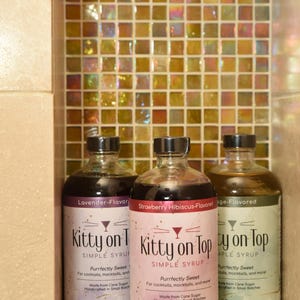 May include: Three glass bottles of flavored simple syrup, labelled "Kitty on Top." Flavours include Lavender, Strawberry Hibiscus, and Sage. Each bottle is 473 ml. The background features a mosaic tile wall.