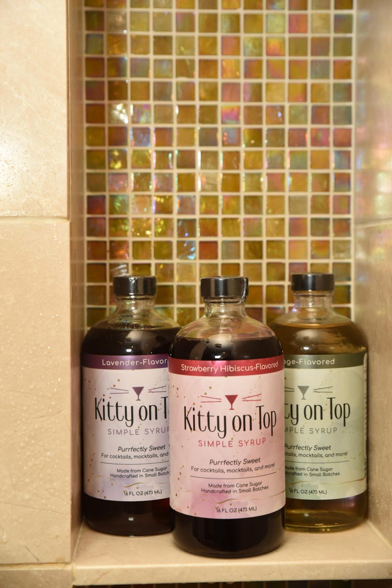 May include: Three glass bottles of flavored simple syrup, labelled "Kitty on Top." Flavours include Lavender, Strawberry Hibiscus, and Sage. Each bottle is 473 ml. The background features a mosaic tile wall.