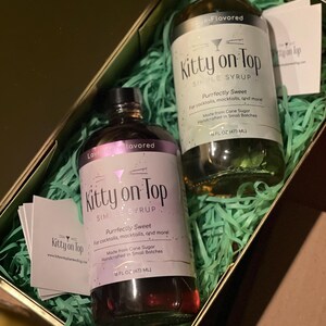 May include: A gift box containing two bottles of "Kitty on Top" simple syrup. One bottle is lavender-flavored, the other sage-flavored. Both bottles are 473 ml and are labeled "Purrfectly Sweet" for cocktails and mocktails. The box is filled with green paper.