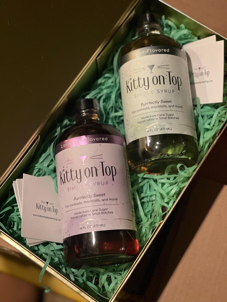May include: A gift box containing two bottles of "Kitty on Top" simple syrup. One bottle is lavender-flavored, the other sage-flavored. Both bottles are 473 ml and are labeled "Purrfectly Sweet" for cocktails and mocktails. The box is filled with green paper.