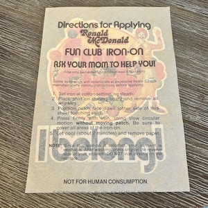 Może przedstawiać: Vintage termotransfer z Ronald McDonald Fun Club z instrukcją. Projekt przedstawia Ronalda McDonalda i tekst "Ask Your Mom to Help You!" na beżowym tle. Zawiera instrukcje dotyczące aplikacji.