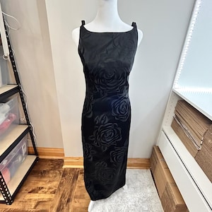 May include: A black, floor-length dress with a floral rose pattern. The dress has a square neckline and thick straps. The fabric appears to be a satin or similar material. The dress is displayed on a white mannequin.