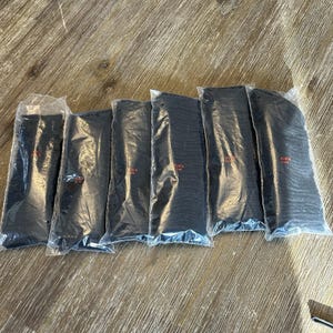 Könnte beinhalten: Sechs einzelne Paare schwarzer Socken, jeweils in einer durchsichtigen Kunststoffhülle versiegelt. Jede Verpackung ist auf der Vorderseite mit dem Text "BLACK 0144" in kleinen roten Buchstaben bedruckt. Die Socken sind in einer Reihe angeordnet.