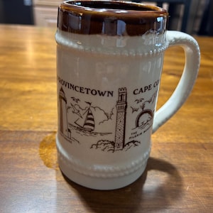 Op de afbeelding: Een crèmekleurige keramische mok met een bruine rand en handvat. De mok heeft bruine illustraties van een vuurtoren, een zeilboot en de tekst "Provincetown Cape Cod". De mok heeft een kralenontwerp bij de basis.