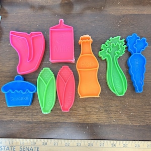 May include: A collection of colorful plastic cookie cutters in various shapes, including a cupcake, corn, milk carton, pop bottle, celery, and carrot. The cutters are in shades of pink, orange, green, and blue.
