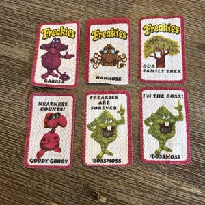 May include: Six vintage "Freakies" patches with cartoon characters and text. Each patch has a white background with a red border. The patches feature characters named Garcle, Hamhose, Bossmoss, and Goody-Goody, with phrases like "Neatness Counts!" and "I'm the Boss!"