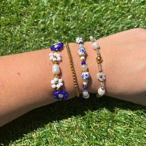 May include: Four beaded bracelets in various designs. One bracelet features blue and white flower-shaped beads, another has a gold chain, and two others have a mix of white, blue, and gold beads. The bracelets are displayed on a person's arm against a green grass background.