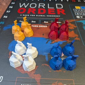 Può includere: Un gioco da tavolo intitolato World Order: A Race for Global Hegemony che mostra una mappa delle Americhe. Sedici piccole pedine di plastica a forma di cupola con bandierine sono disposte in quattro gruppi da quattro, di colore giallo, rosso, bianco e blu.