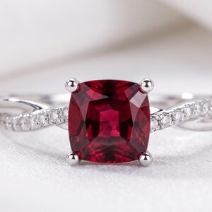 May include: A ring featuring a central cushion-cut deep red gemstone held by four prongs. The band is crafted from a silver-toned metal and features a twisted design on both sides, accented with small, clear, round-cut stones set along the shank.