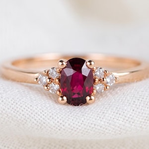 May include: A rose gold ring featuring a central oval-cut deep red ruby gemstone. The ruby is flanked on each side by a cluster of three small, round, clear white diamonds set into the band, creating a delicate and balanced design.