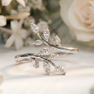 May include: A silver-toned bypass ring featuring two delicate, branching stems adorned with multiple marquise-cut clear stones. The design mimics a natural leaf motif, with the stems curving around each other to form an open, adjustable band.