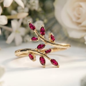 May include: A gold-colored ring with a leaf design, adorned with small, oval-shaped red gemstones. The ring is set against a backdrop of white flowers, creating a delicate and elegant aesthetic. The ring's design resembles a branch with leaves.