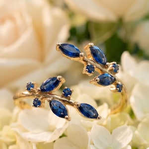 May include: A gold ring featuring a leaf design, embellished with blue gemstones. The ring is displayed on a bed of white flowers, offering a contrast of colours and textures. The gemstones are oval-shaped, adding a touch of sophistication.