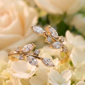 May include: Two gold-colored rings with a leaf design, each adorned with multiple clear, sparkling gemstones. The rings are set against a backdrop of white flowers and soft, blurred cream-colored roses, creating a romantic aesthetic.