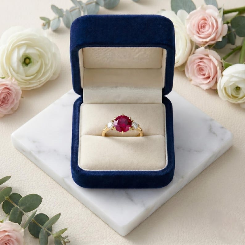 Pu&ograve; includere: Un anello in oro con una grande pietra centrale di rubino rosso a taglio rotondo, affiancata da due diamanti bianchi pi&ugrave; piccoli a taglio rotondo. L'anello &egrave; esposto all'interno di un portagioie in velluto blu navy con interno color crema.