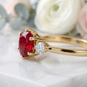 Pu&ograve; includere: Un anello in oro con una grande pietra preziosa rosso vivo a taglio ovale come pietra centrale, affiancata da un diamante bianco chiaro a taglio rotondo su ciascun lato. La fascia &egrave; in oro giallo lucido, con un classico design a tre pietre.