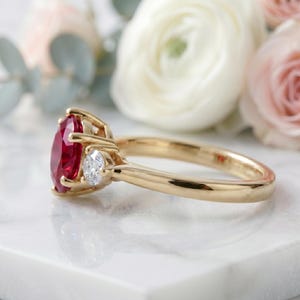 Pu&ograve; includere: Un anello in oro con un grande rubino rosso taglio ovale come pietra centrale, affiancato da un diamante bianco taglio rotondo pi&ugrave; piccolo su ciascun lato. La fascia &egrave; realizzata in oro giallo lucido con un design classico a tre pietre.