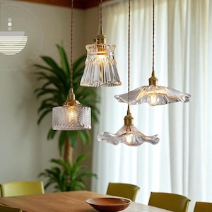 May include: Three glass pendant lights with brass fittings hang above a wooden table. The glass shades have different designs, including ribbed, textured, and scalloped edges. The lights are suspended from brown cords.