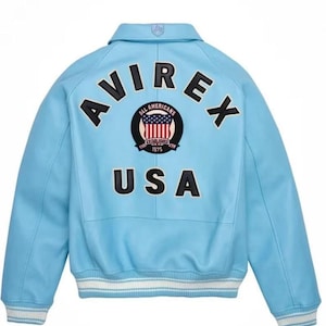 Puede incluir: Chaqueta bomber azul claro con la palabra "AVIREX" en letras negras en la espalda. Un parche con las palabras "ALL AMERICANS ESTABLISHED 1975" rodea un gr&aacute;fico de la bandera estadounidense. La palabra "USA" est&aacute; debajo del parche.