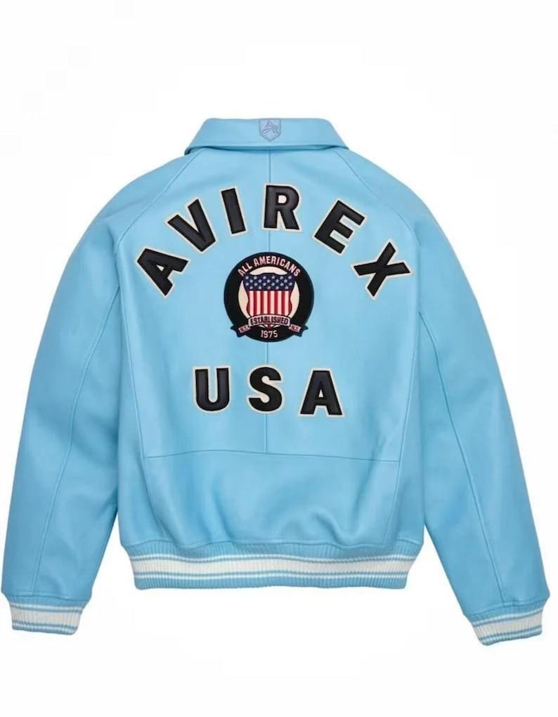Puede incluir: Chaqueta bomber azul claro con la palabra "AVIREX" en letras negras en la espalda. Un parche con las palabras "ALL AMERICANS ESTABLISHED 1975" rodea un gr&aacute;fico de la bandera estadounidense. La palabra "USA" est&aacute; debajo del parche.