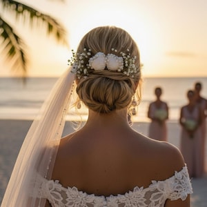Op de afbeelding: Een bruid met blond haar in een lage knot, versierd met haaraccessoires van zeeschelpen en gipskruid. Ze draagt een witte kanten off-the-shoulder trouwjurk en een doorschijnende sluier. Het strand en de bruidsmeisjes zijn op de achtergrond.