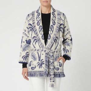 Op de afbeelding: Crèmekleurig vest met een blauw paisley- en palmbomenpatroon. Het vest heeft een sjaalkraag, een bijpassende riem en franjes. De mouwen zijn lang en de manchetten zijn marineblauw. Het vest wordt gedragen over een zwarte top en een witte broek.