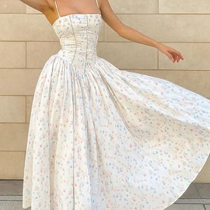 Puede incluir: Un vestido blanco con estampado floral con un corpiño ajustado y una falda fluida. El vestido presenta tirantes finos y un corpiño estilo corsé. La tela está adornada con pequeños estampados florales azules, rosas y amarillos. El vestido es adecuado para ocasiones especiales.